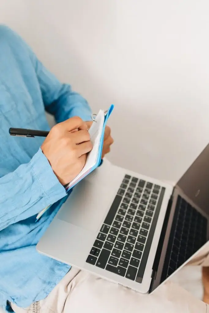 Close-up of an adult writing notes on a laptop in a home setting.